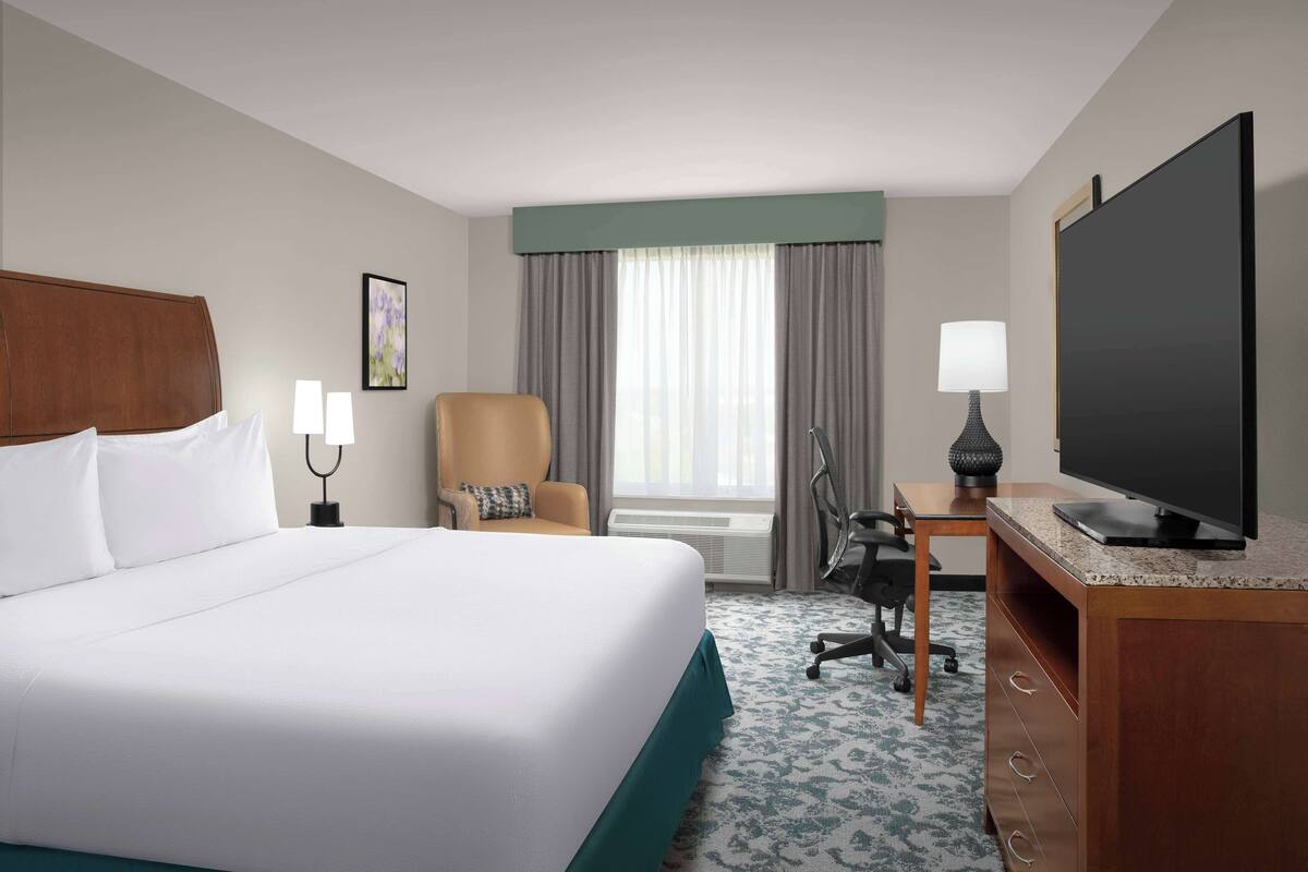A hotel room with a large bed, a desk with a television, a chair, and a window with curtains.