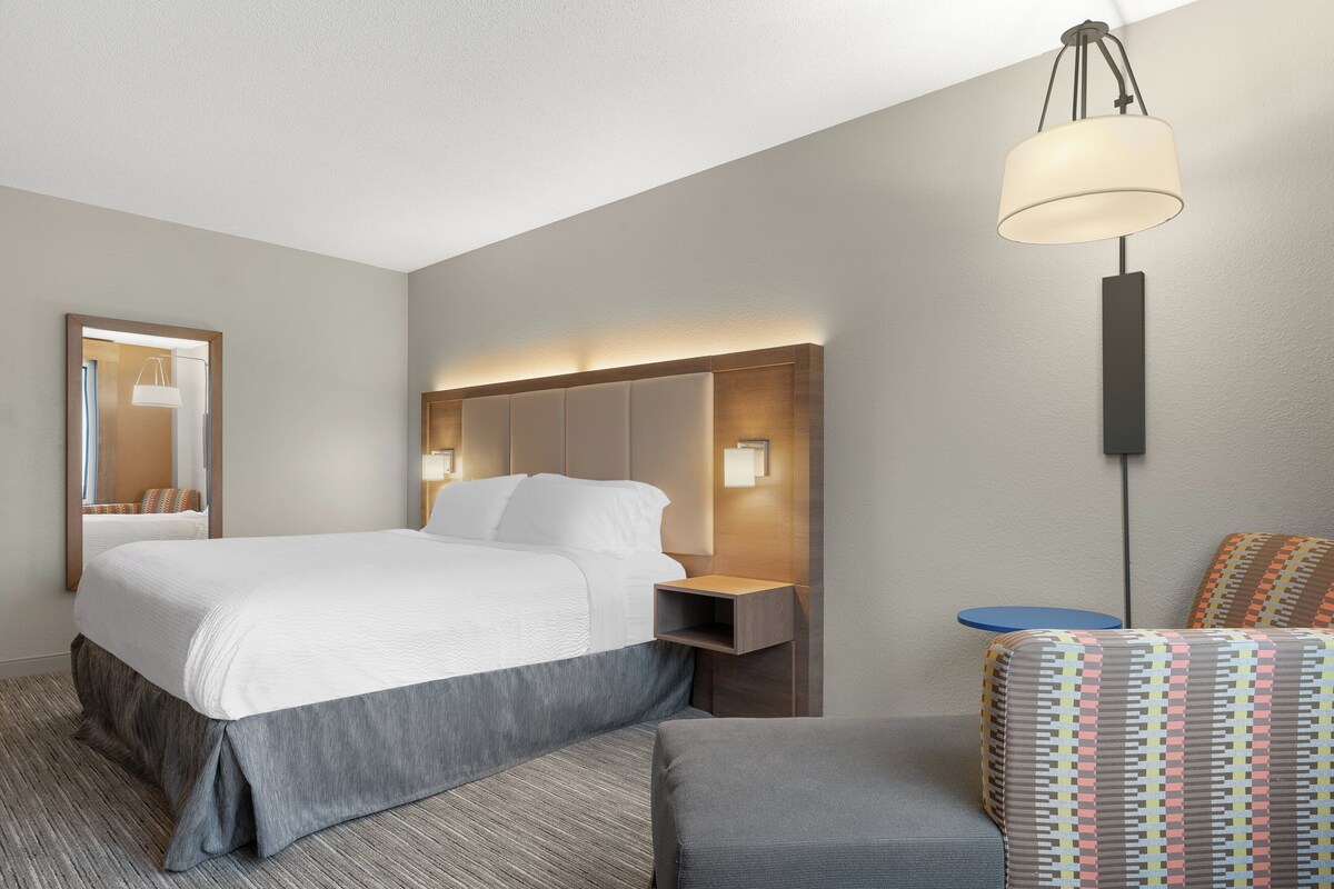 Ein Hotelzimmer mit einem großen Bett, Nachttischen, einem Spiegel, einer Hängelampe und einem gemusterten Sessel.
