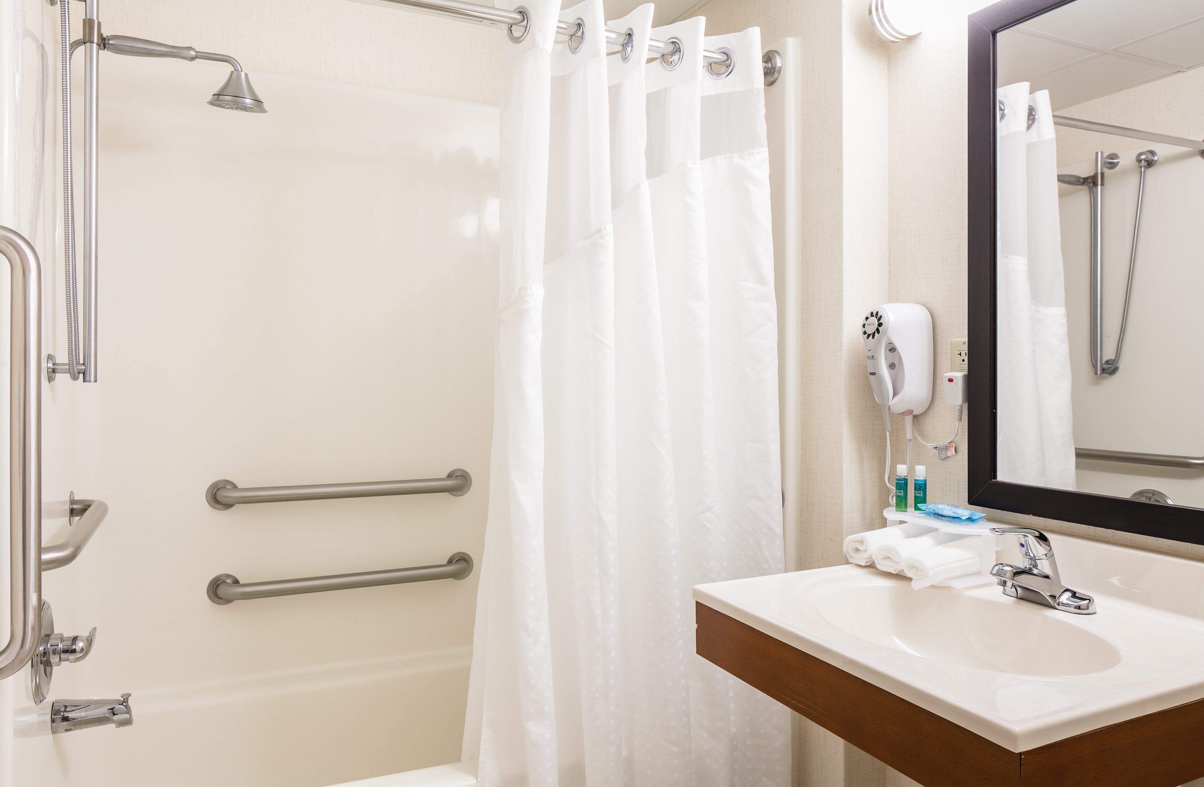 A bathroom with a shower, grab bars, a sink, and a mirror.