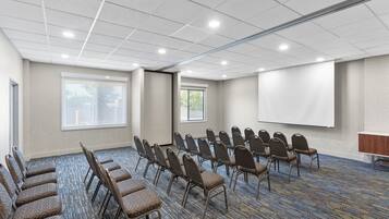 Un salón de conferencias con filas de sillas, una pantalla de proyector y ventanas amplias.
