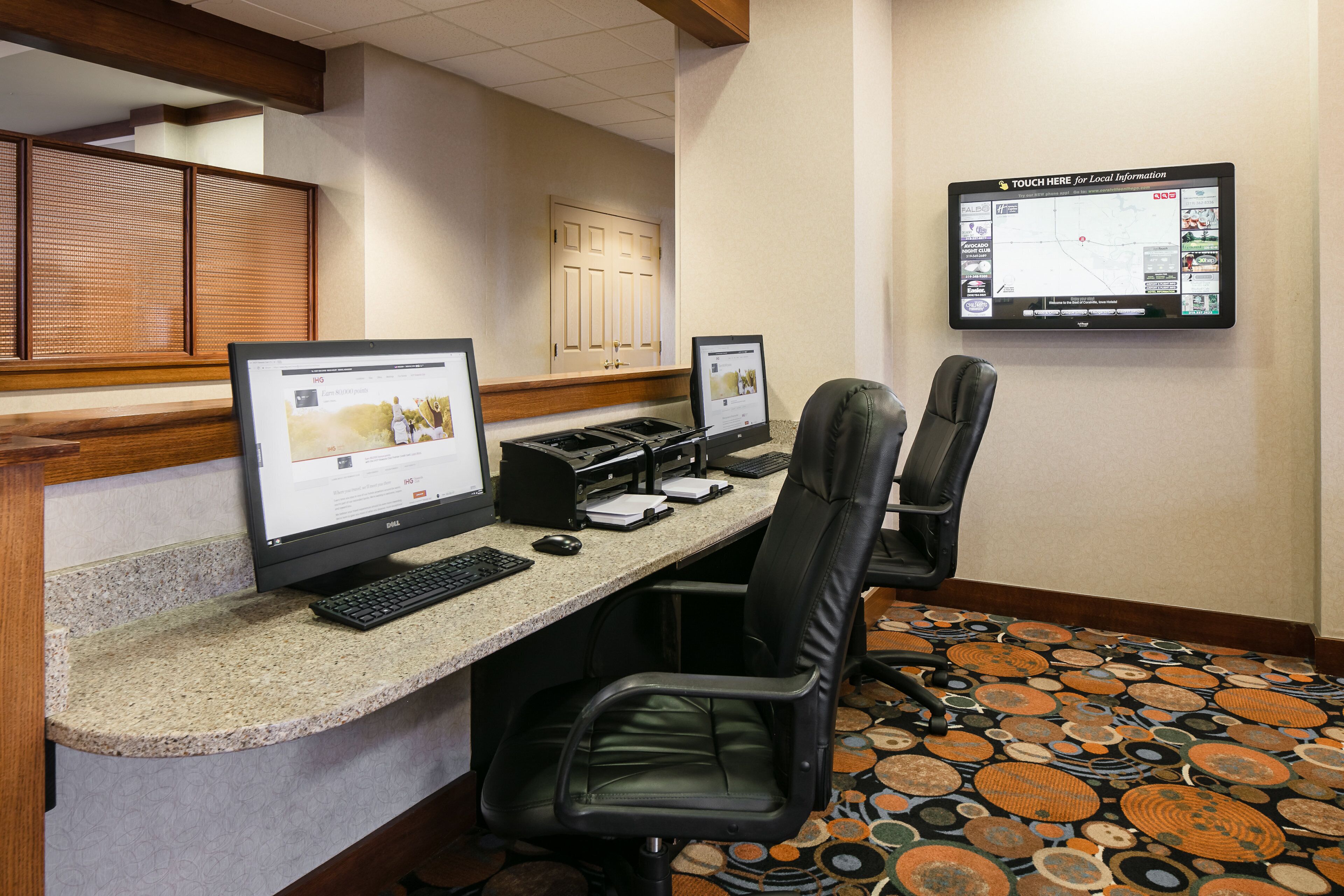 A hotel reception area with two desktop computers, a printer, and a large screen displaying local information.