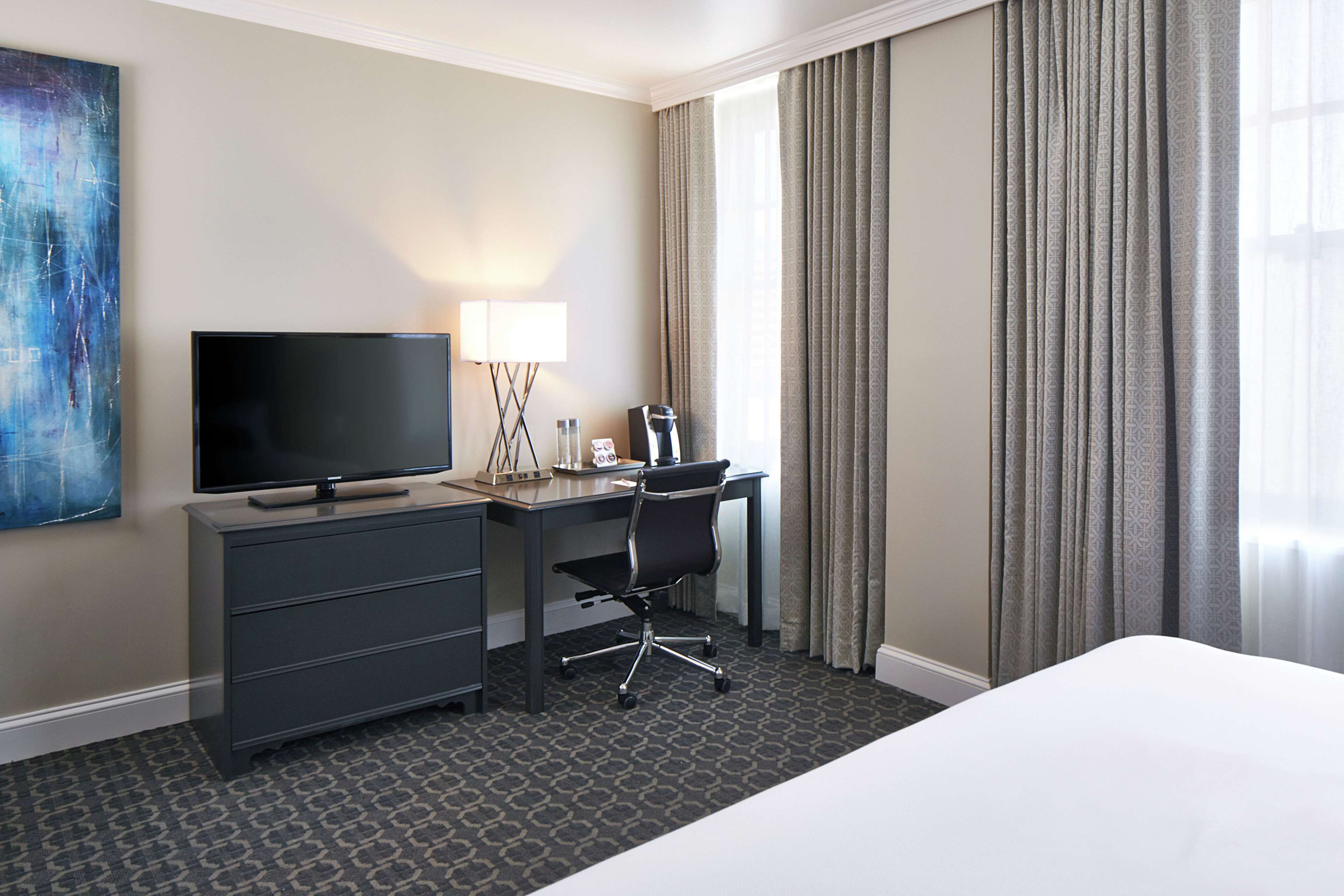 A hotel room with a bed, a desk with a television, a chair, and a lamp.