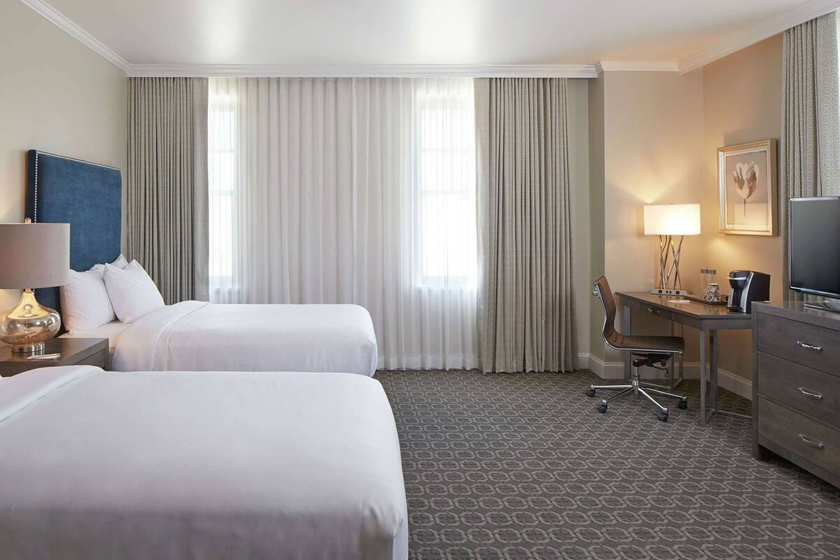 A hotel room with two beds, a desk, a chair, a television, and a lamp.