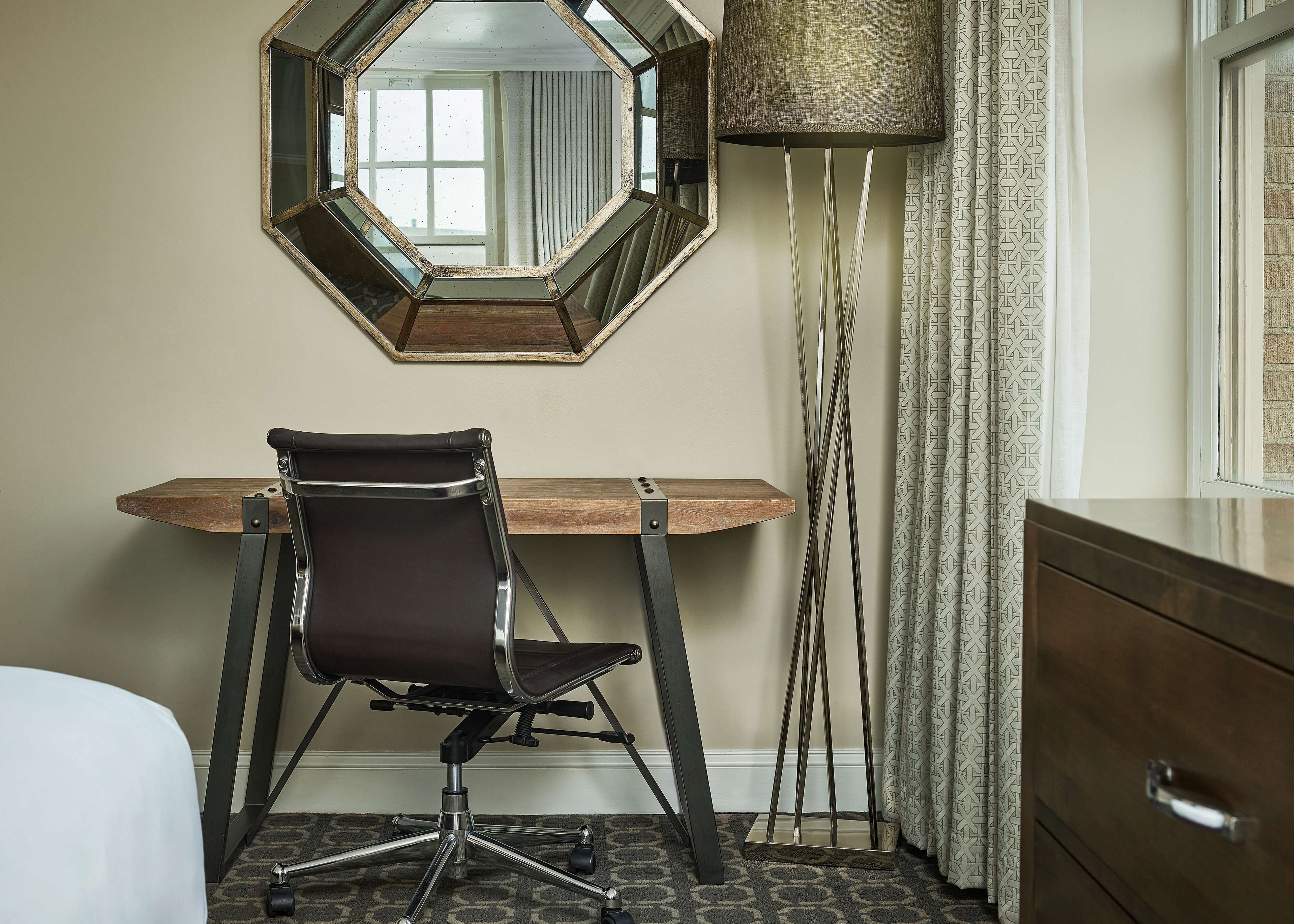 Habitación de hotel con un escritorio de madera, una silla de oficina negra, un espejo grande, una lámpara de pie y un aparador.