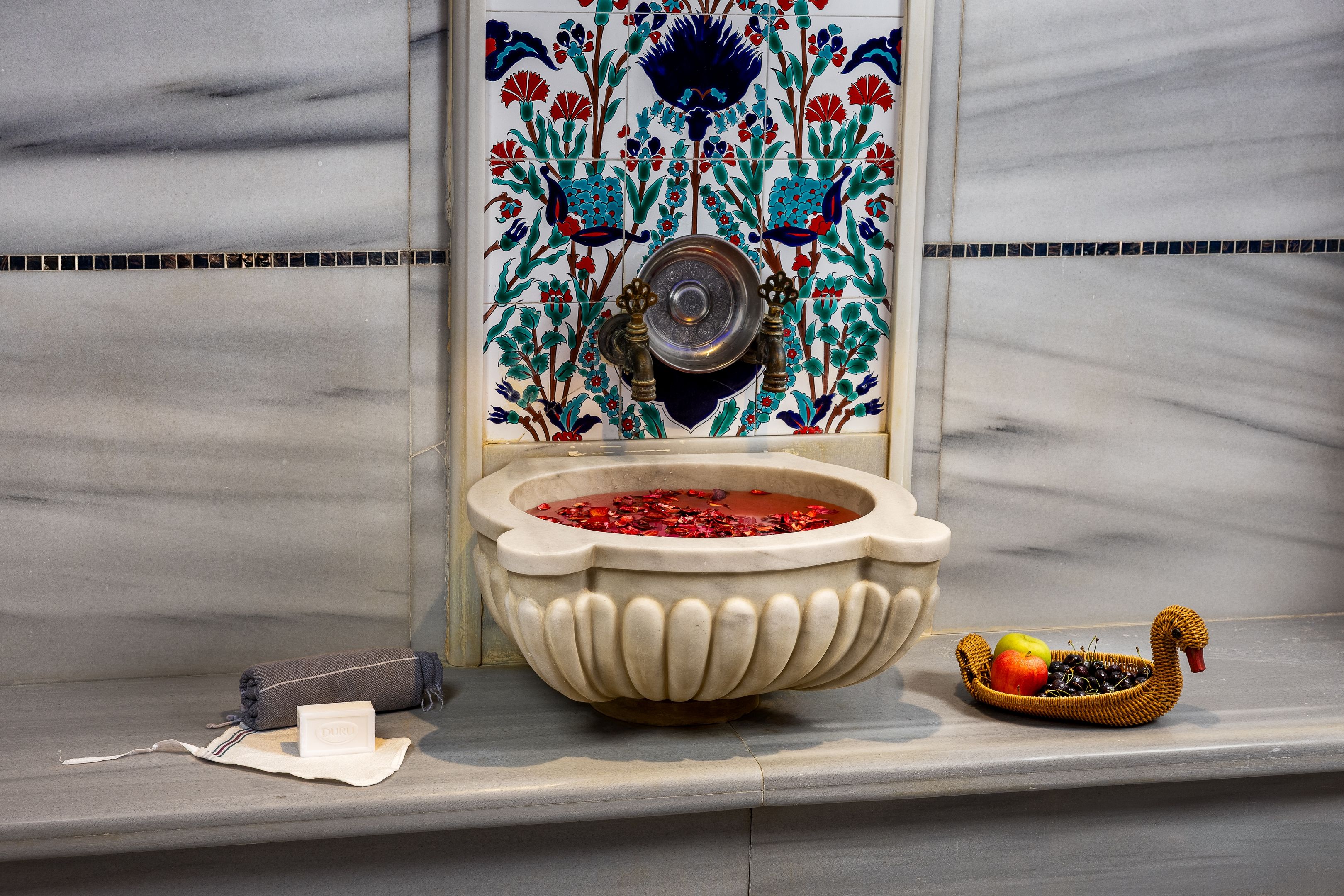 A bathroom with a decorative tile wall, a stone basin with water, and a wicker basket with fruits.