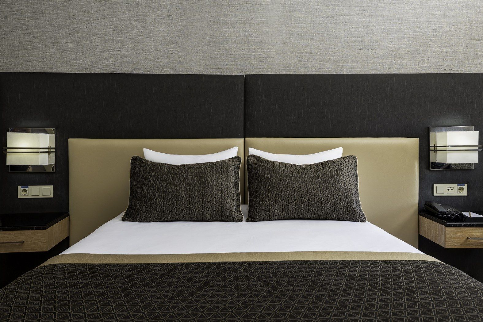 A hotel room with a large bed, two pillows, a headboard, bedside tables, and wall-mounted lamps.