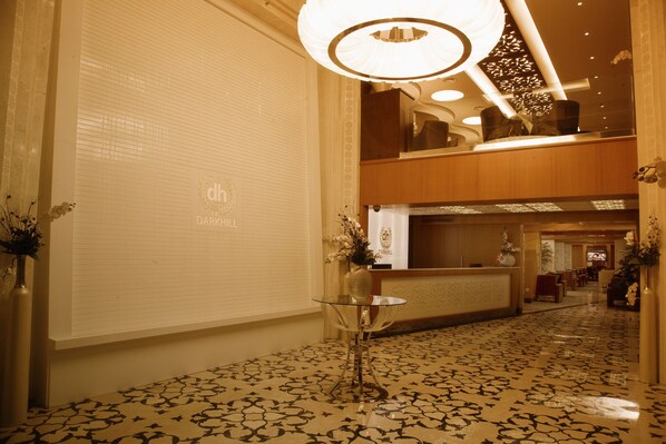 Un hall d’hôtel comprenant un comptoir d’accueil, une table en verre ornée d’un vase, un revêtement de sol à motifs et un lustre.