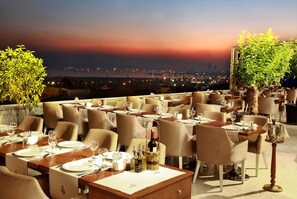 A rooftop restaurant with tables set for dining, a view of the city skyline at dusk, and a large tree in the foreground. - Darkhill Hotel (Istanbul)