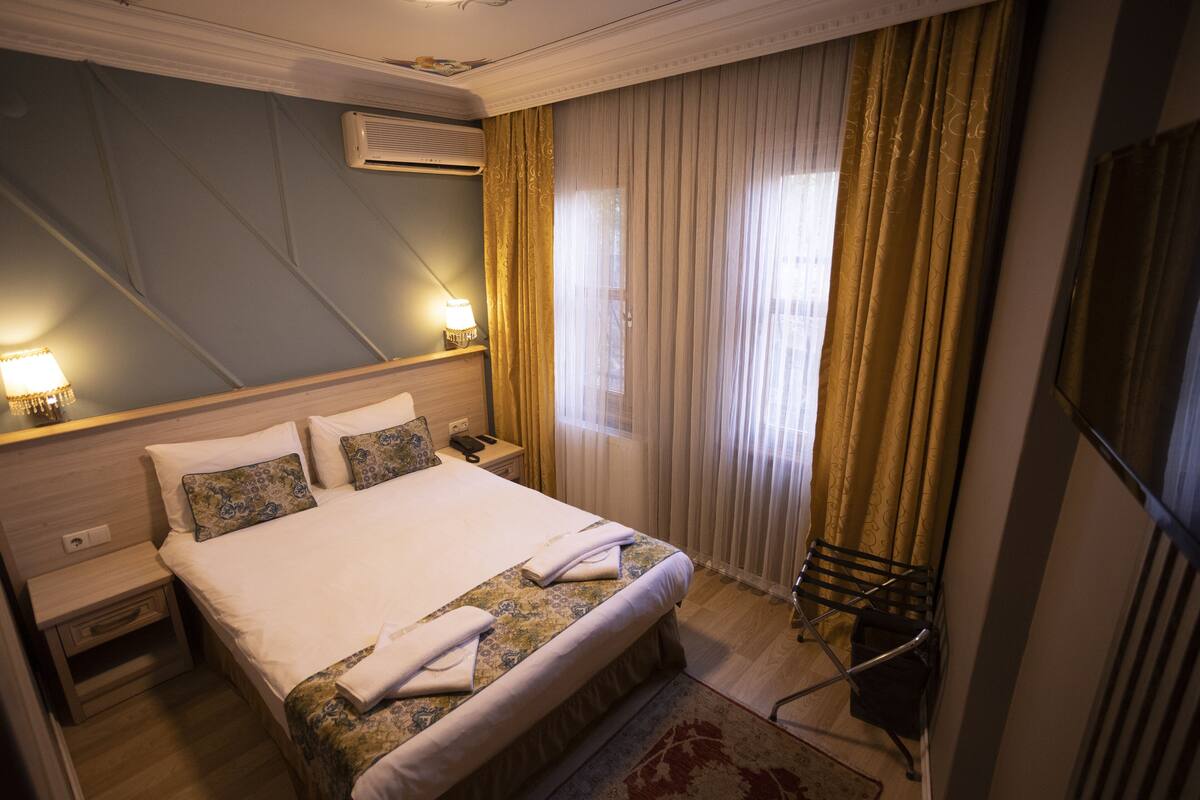 A hotel room with a large bed, two bedside tables, a lamp, a wall-mounted air conditioner, and a window with curtains.