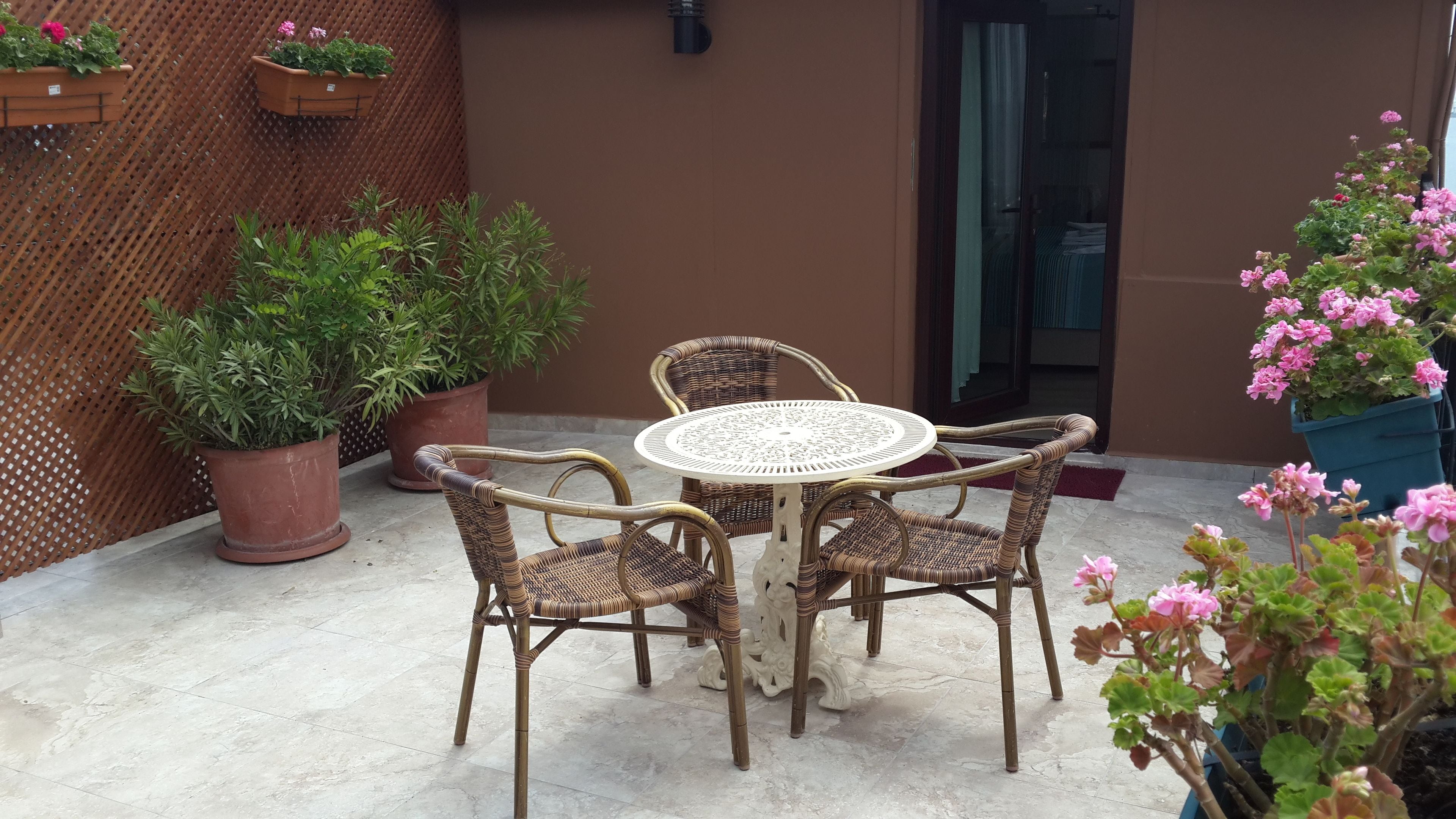 A patio with a round table and four wicker chairs, surrounded by potted plants and a wooden lattice wall.