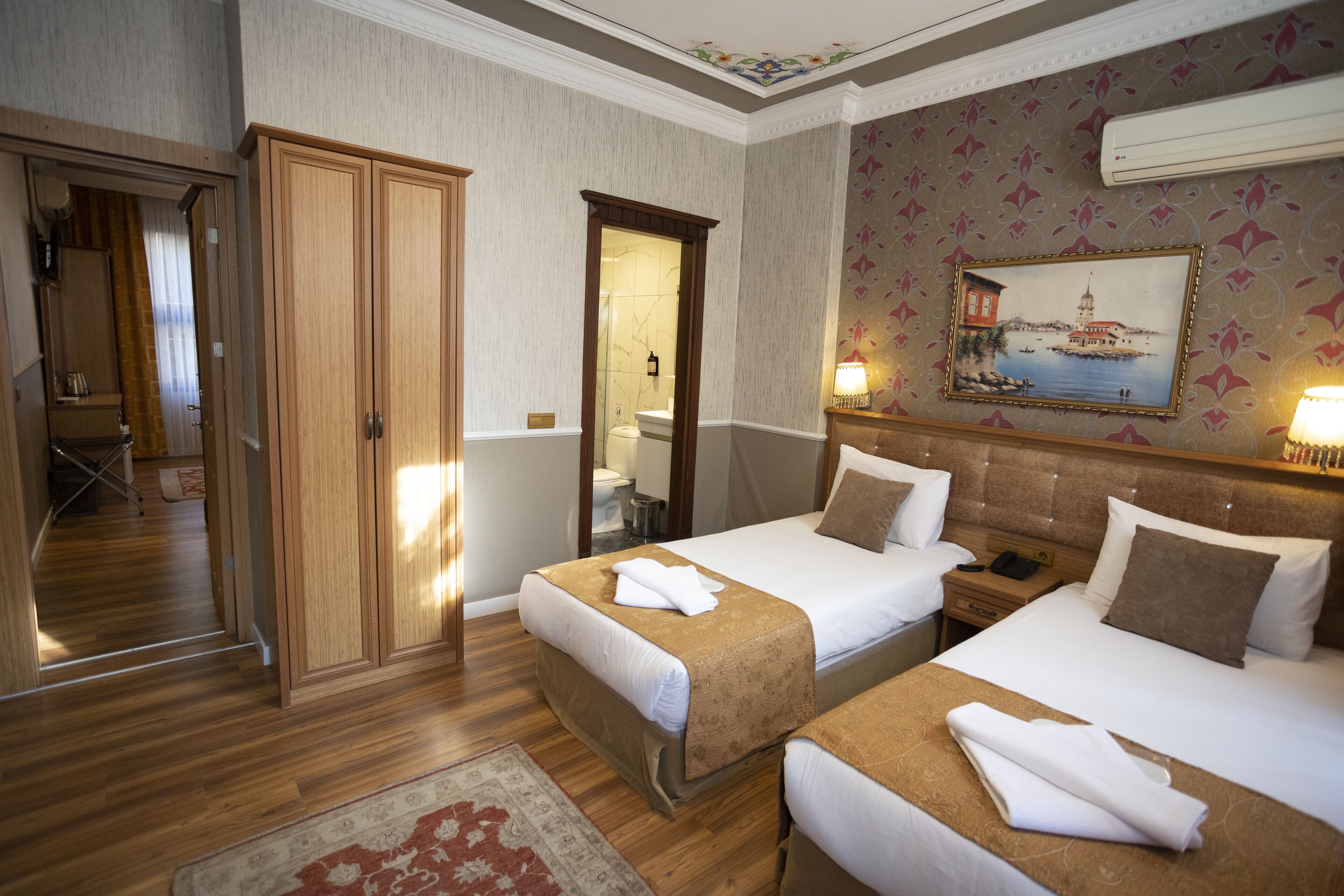 A hotel room with two beds, a wooden wardrobe, a bathroom, and a painting on the wall.