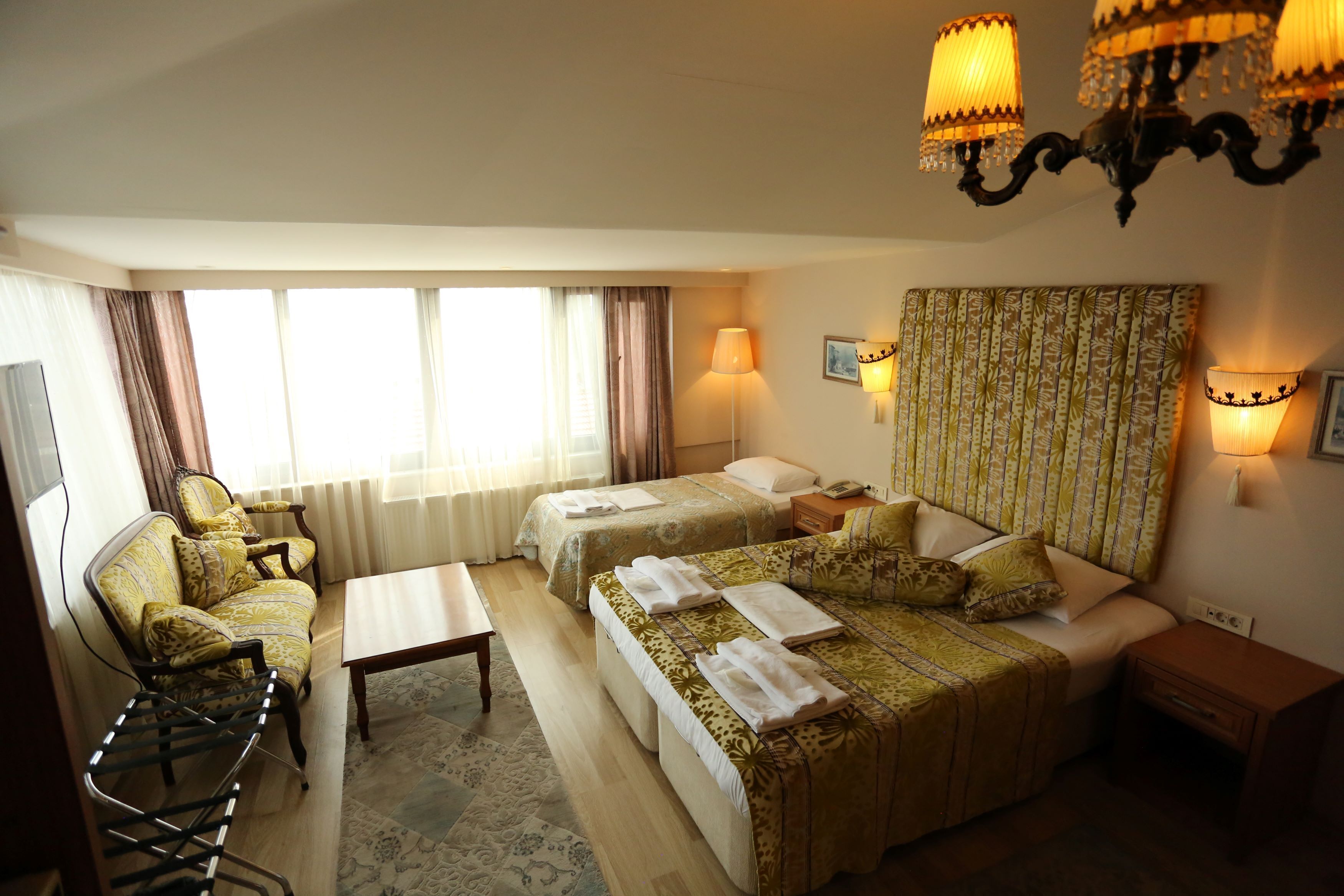 A hotel room with two beds, a sofa, a table, and a chair. There are lamps on the walls and a chandelier.