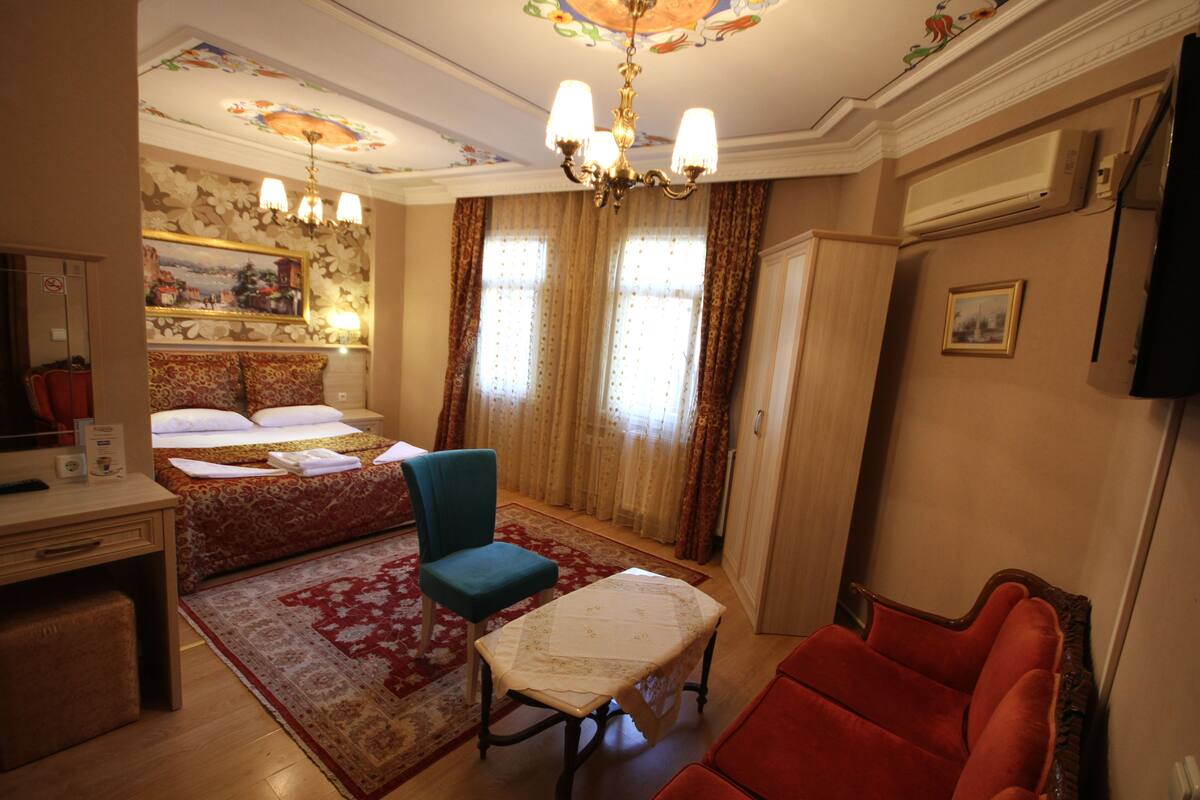 A hotel room with a bed, a desk, a chair, a table, a red armchair, a chandelier, curtains, and a painting on the wall.