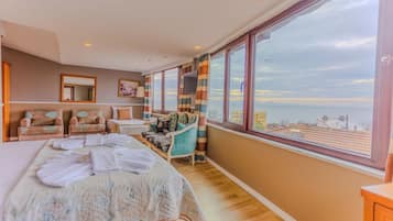 Un dormitorio amplio con una cama grande, un área de descanso con sillas, un televisor y un ventanal con vistas al mar.