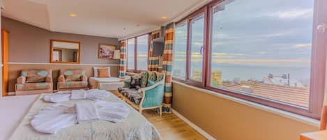A spacious bedroom with a large bed, a seating area with chairs, a television, and a large window offering a view of the sea.