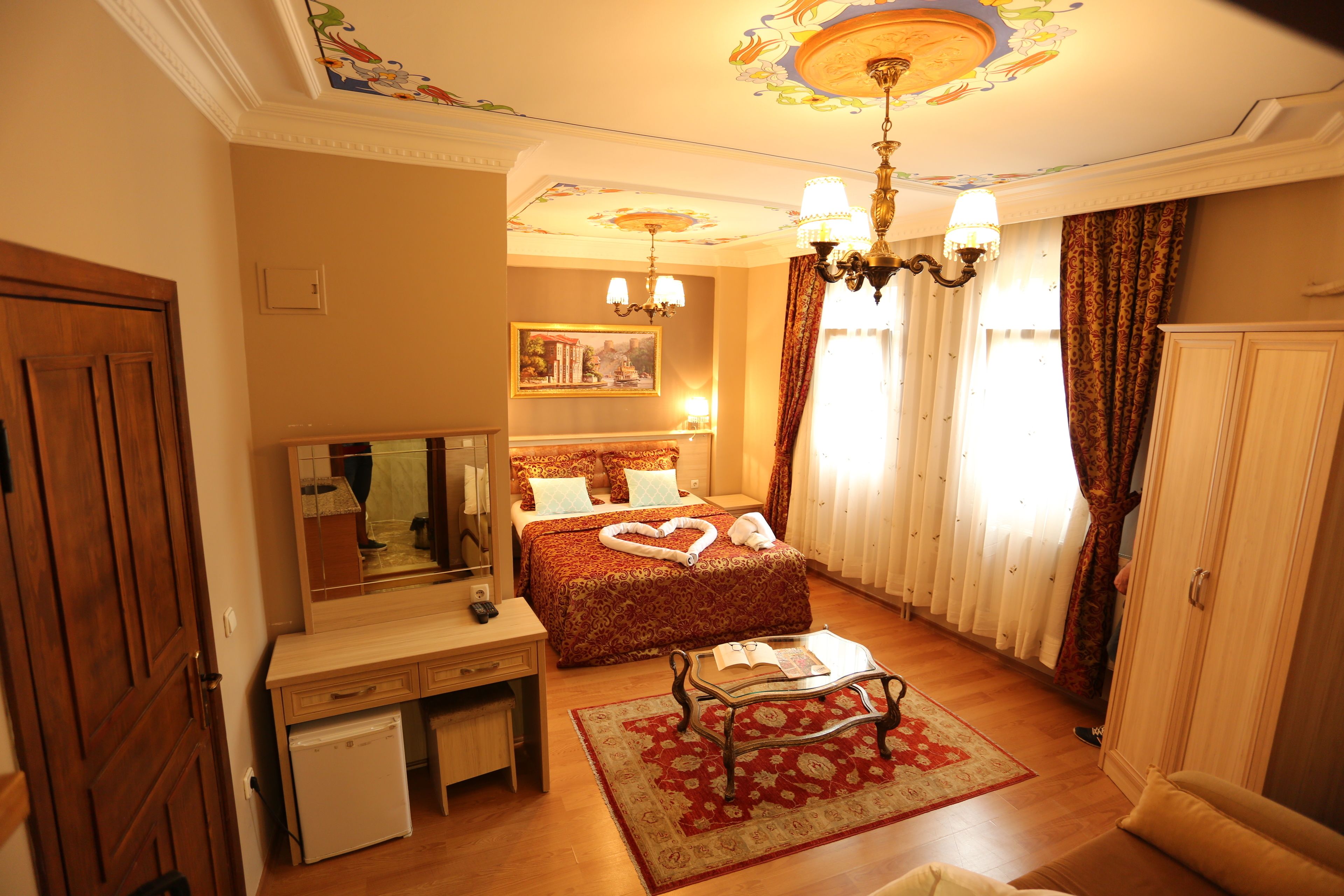 A hotel room with a wooden desk, a bed with a heart-shaped towel, a chandelier, and a large mirror.
