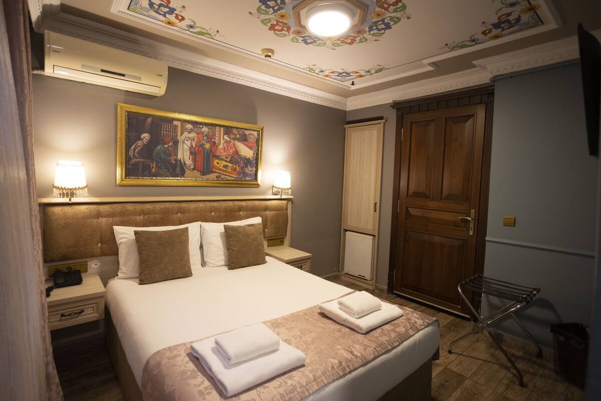A hotel room with a large bed, two bedside tables, a wooden door, and a ceiling with floral patterns.