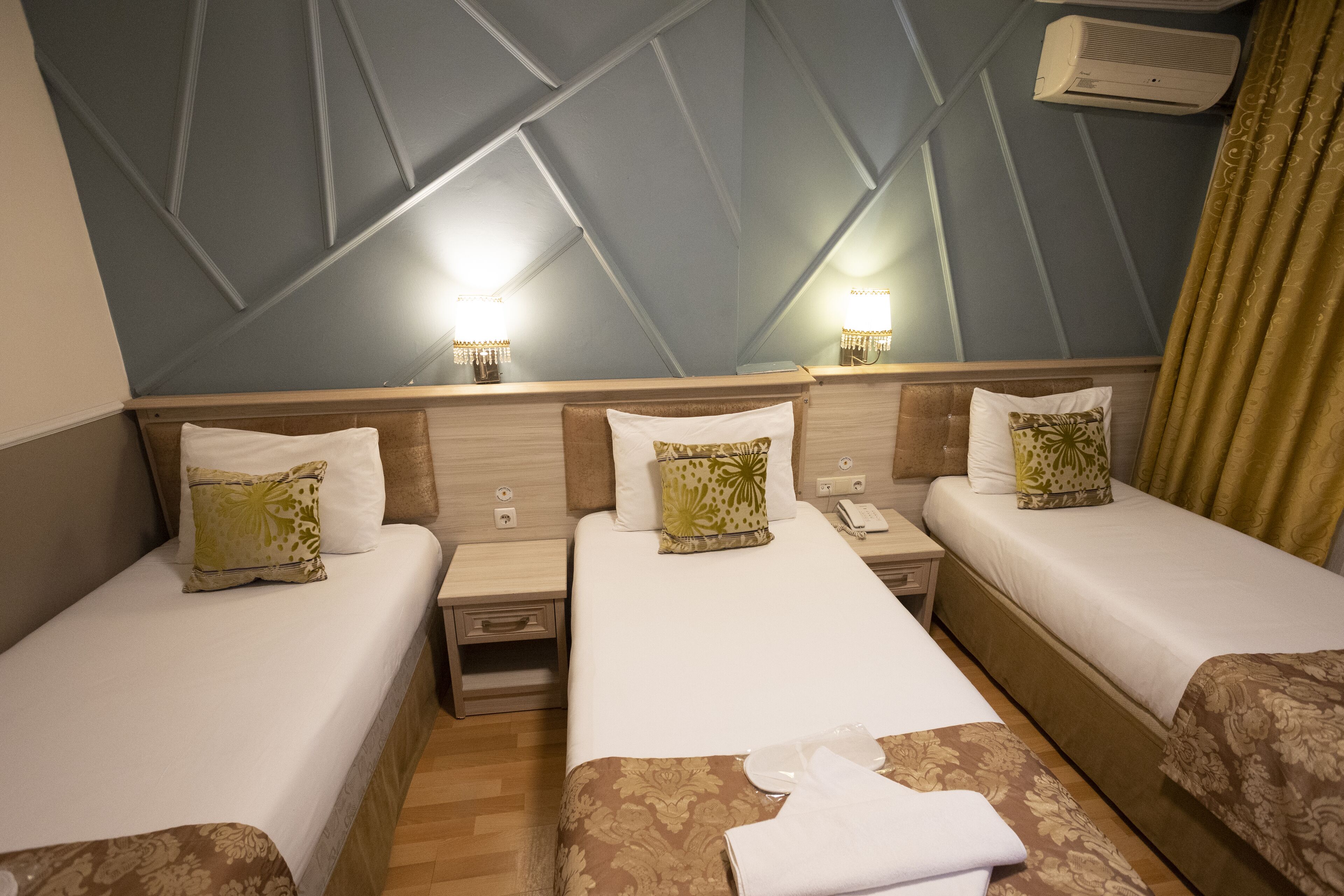 Habitación de hotel con tres camas, cada una con una almohada y una mesita de noche. Hay un aire acondicionado de pared y dos apliques de luz.