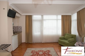 A room with a green sofa, a small kitchen area with a refrigerator, a television mounted on the wall, and a large window with curtains.