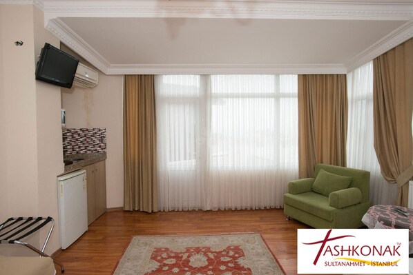 A room with a green sofa, a small kitchen area with a refrigerator, a television mounted on the wall, and a large window with curtains. - Tashkonak Hotel (Istanbul)