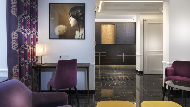 Un moderno hall de hotel con asientos morados y amarillos, un retrato enmarcado en la pared y una recepción elegante.