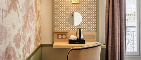 A hotel room with a vanity area, a mirror, a small shelf with a lamp, and a chair.