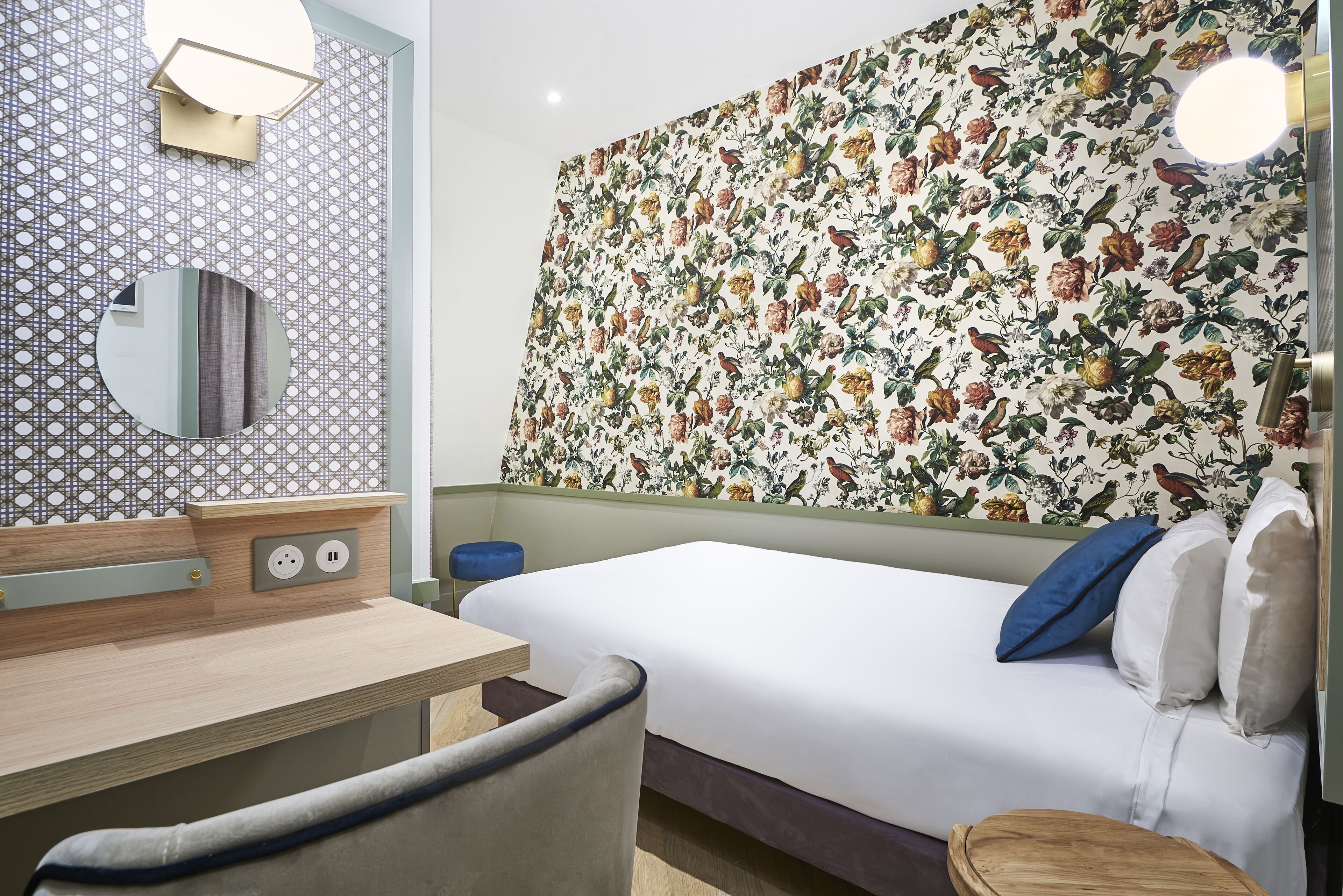 A hotel room with a bed, a desk, a chair, a mirror, and a wall with a floral pattern.