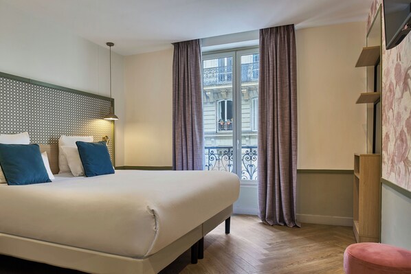 A hotel room with a large bed, a headboard with a patterned design, a window with curtains, and a wooden floor.