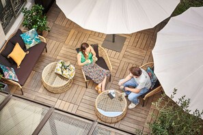 Een terras buiten met rieten meubilair, een witte parasol en plantenpotten.