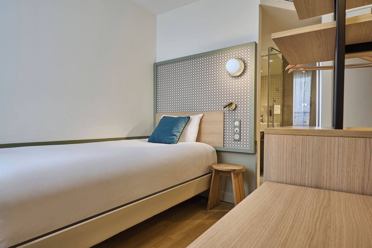 Een moderne hotelkamer met een bed, een klein krukje, een houten hoofdbord en een spiegel.