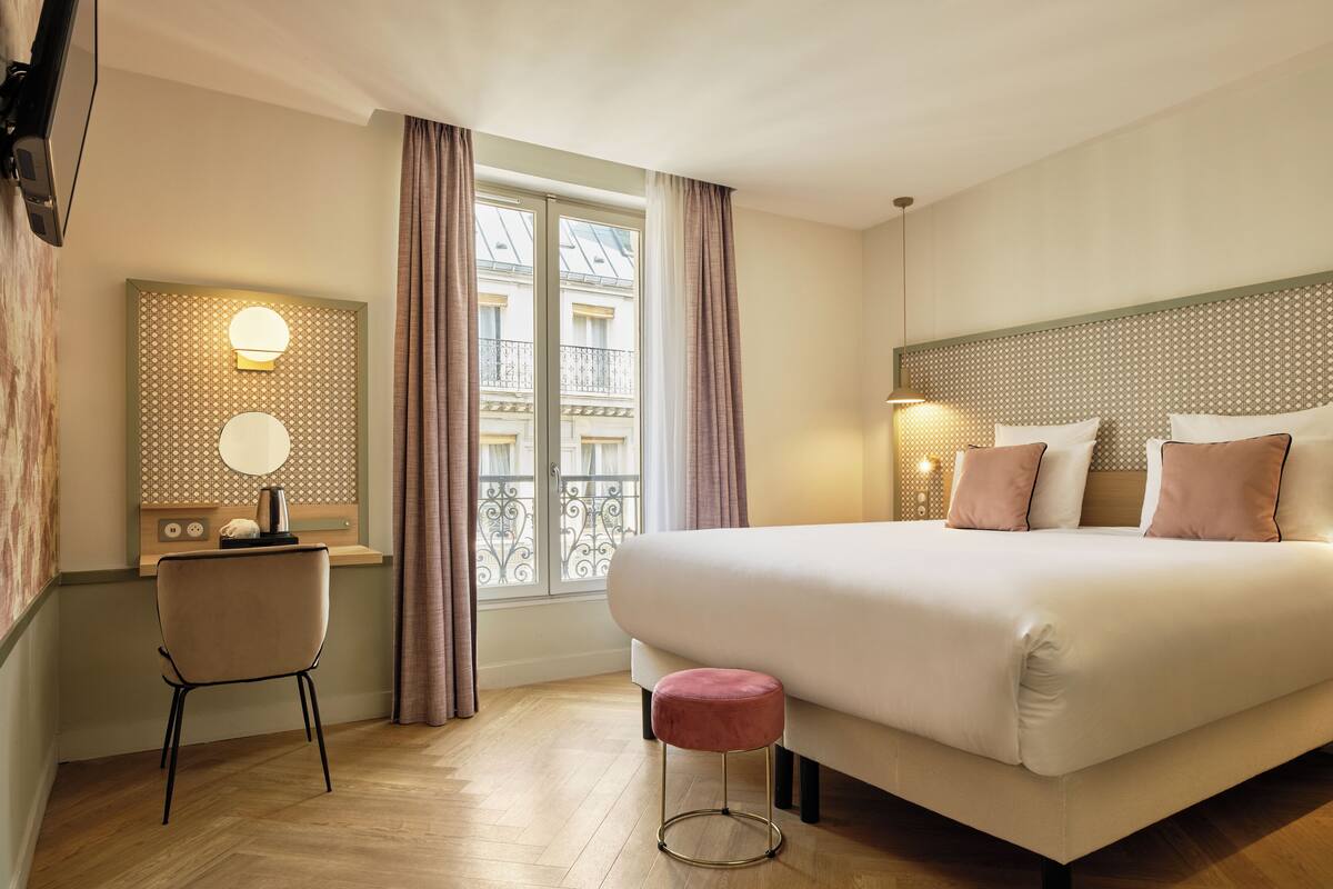 Een hotelkamer met een groot bed, een bureau met stoel, een klein krukje en een raam met gordijnen.
