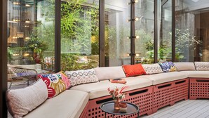 Un espace extérieur moderne aménagé avec un banc rembourré, une petite table ornée d’un vase et d’une tasse, et offrant une vue sur la verdure grâce à de grandes fenêtres.