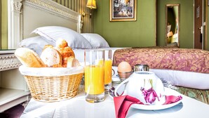 A neatly made bed with a floral pillow, a basket of bread, two glasses of orange juice, a plate with eggs, and a teapot on a table. - Hotel Regence Paris (Paris)