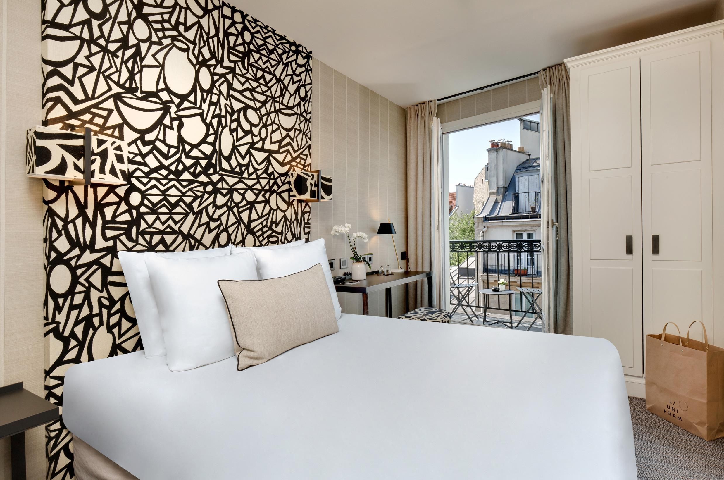 A hotel room with a bed, a patterned wall, a desk with a lamp, and a view of the city through a balcony.