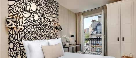 A hotel room with a bed, a patterned wall, a desk with a lamp, and a view of the city through a balcony.