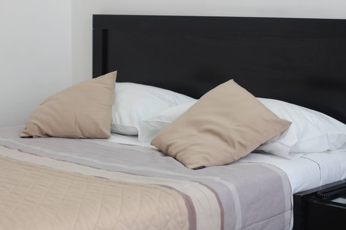 Een net opgemaakt bed met een zwart hoofdbord, beige en witte kussens en een beige deken.