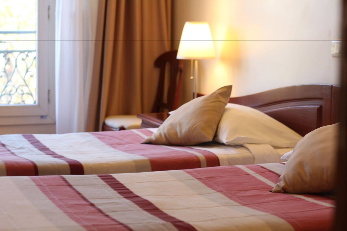 Een hotelkamer met twee bedden, een raam met gordijnen, een bedlampje en een houten hoofdeinde.