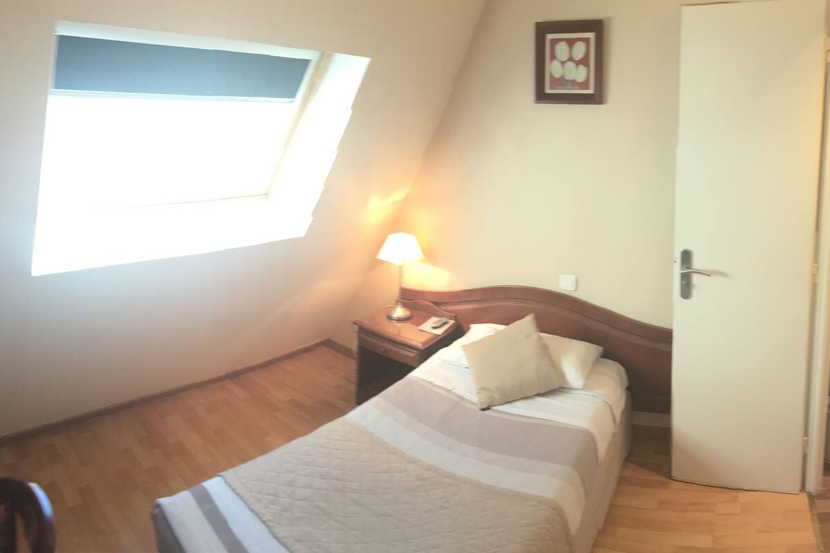 Een kleine, goed verlichte slaapkamer met een enkel bed, een nachtkastje, een lamp en een deur.