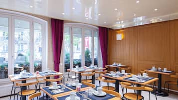 Un comedor de hotel con varias mesas dispuestas para los huéspedes, ventanas amplias con cortinas y vistas al exterior.