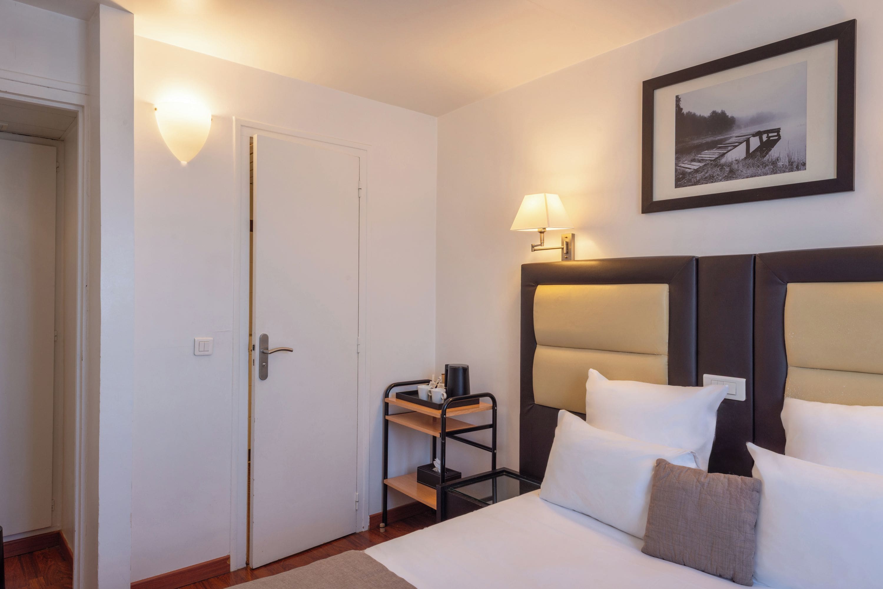 A hotel room with a bed, bedside table, lamp, and a framed picture on the wall.