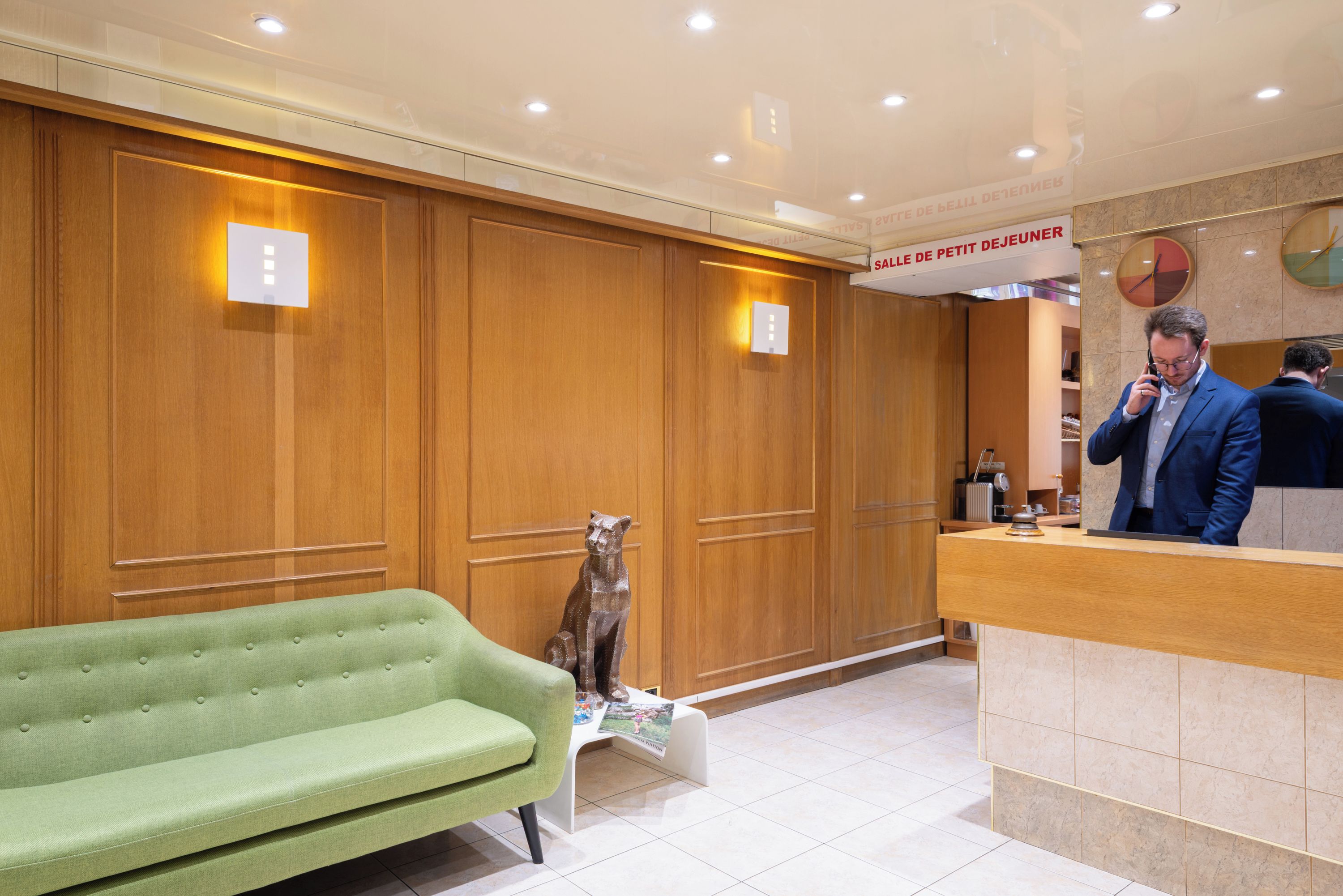 A hotel reception area with a green sofa, wooden paneling, a receptionist desk, and a clock on the wall.