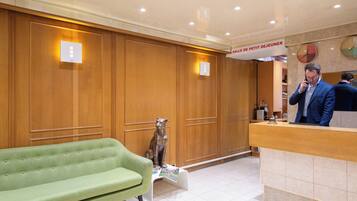 A hotel reception area with a green sofa, wooden paneling, a receptionist desk, and a clock on the wall.