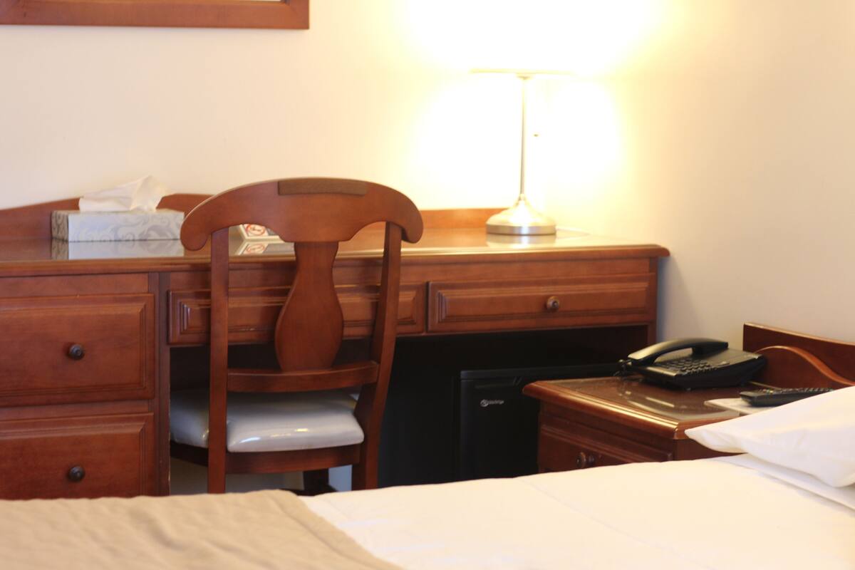 Een hotelkamer met een bed, een houten bureau, een stoel, een lamp, een telefoon en een nachtkastje.