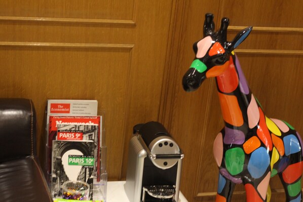 Un mur recouvert de panneaux de bois, un fauteuil en cuir noir, une sculpture colorée représentant une girafe, une machine à café noire et un livre intitulé « The Economist », avec une couverture de la 9e édition de Paris.