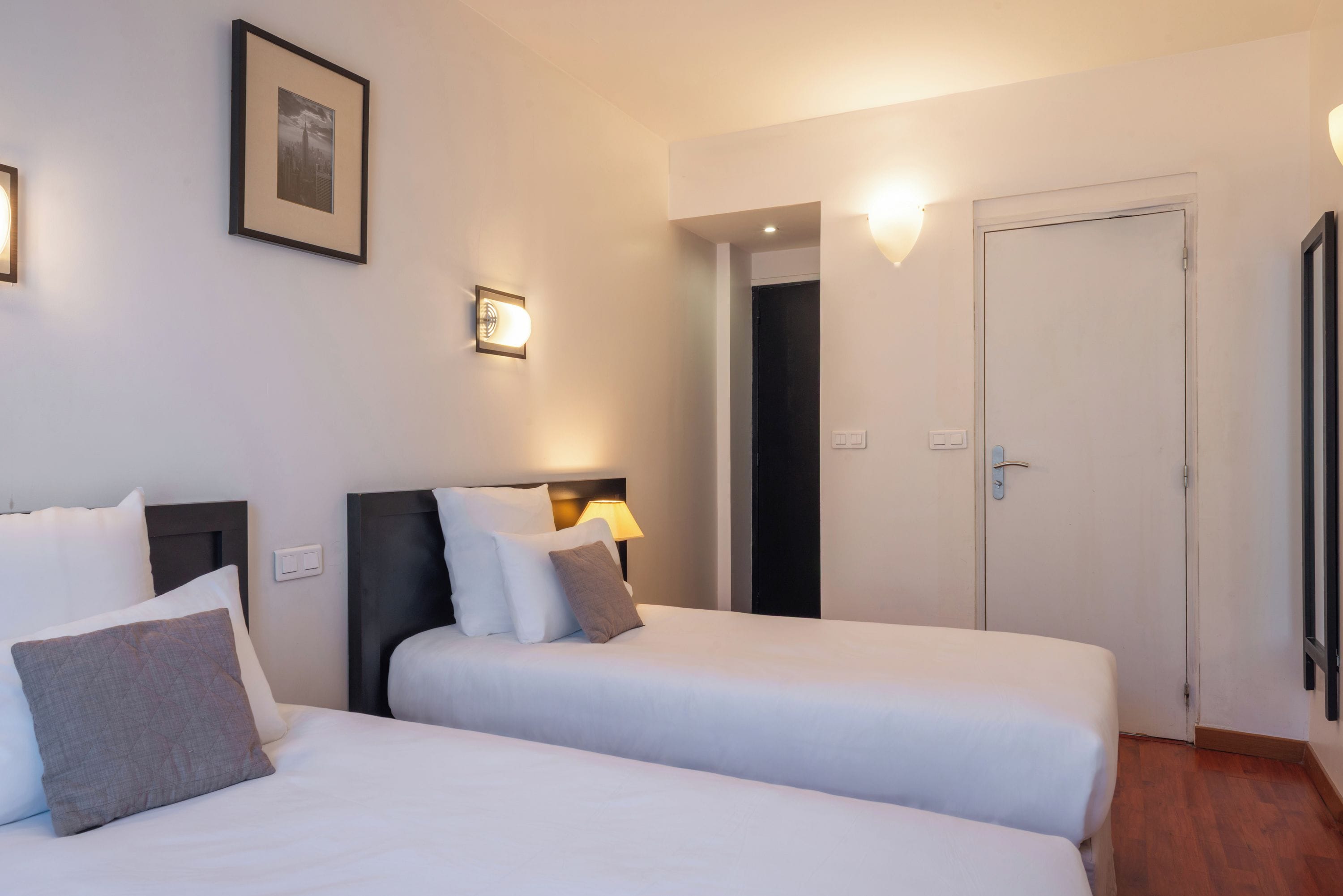 A hotel room with two beds, a framed picture on the wall, a door, and a light fixture.