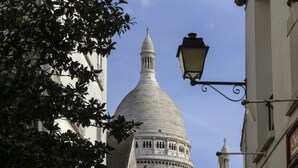 Un bâtiment en forme de dôme, surmonté d’une flèche centrale, entouré de verdure et d’un lampadaire.