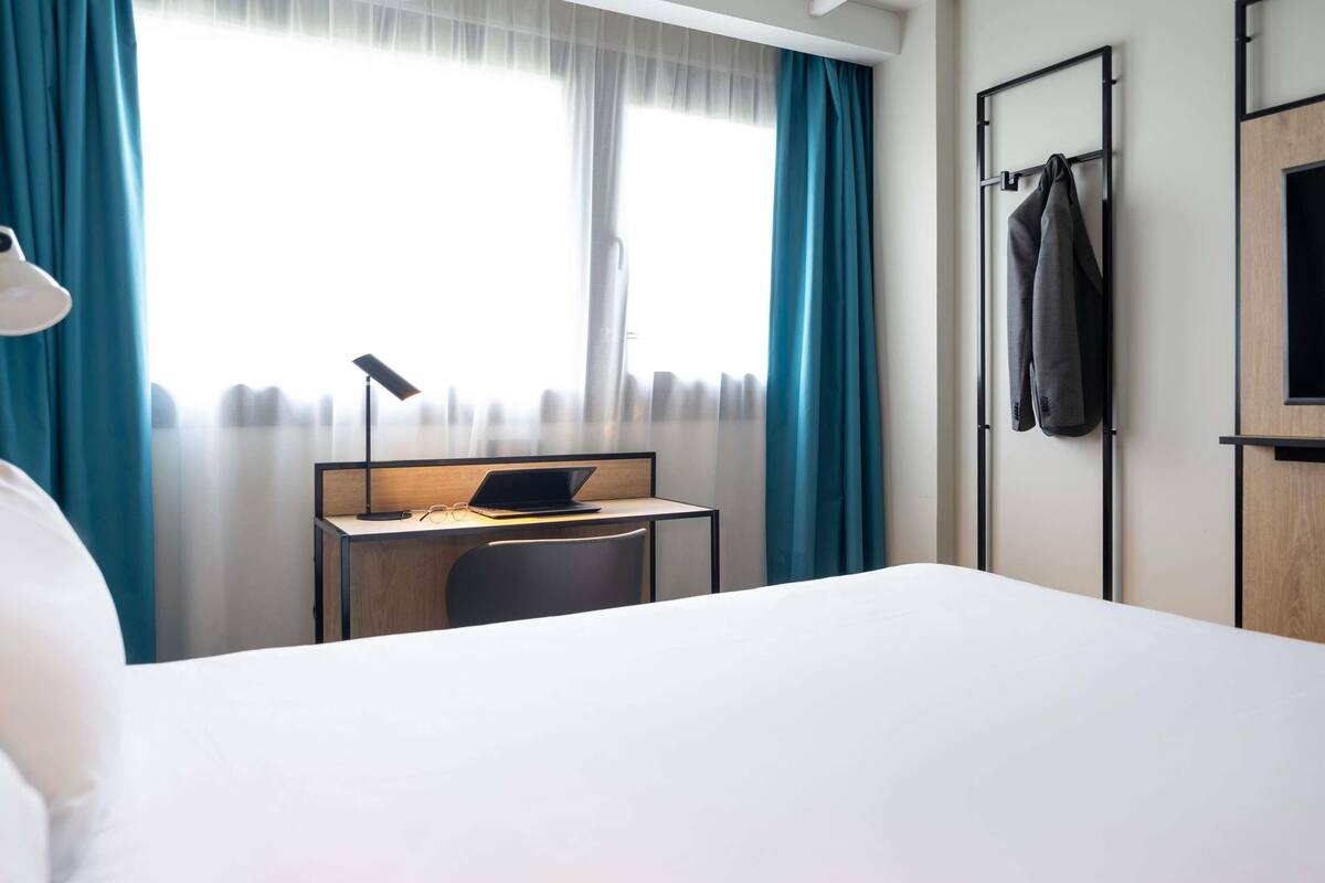 Hotelkamer met een bed, bureau met een laptop, stoel, lamp, kapstok met jas en raam met gordijnen.