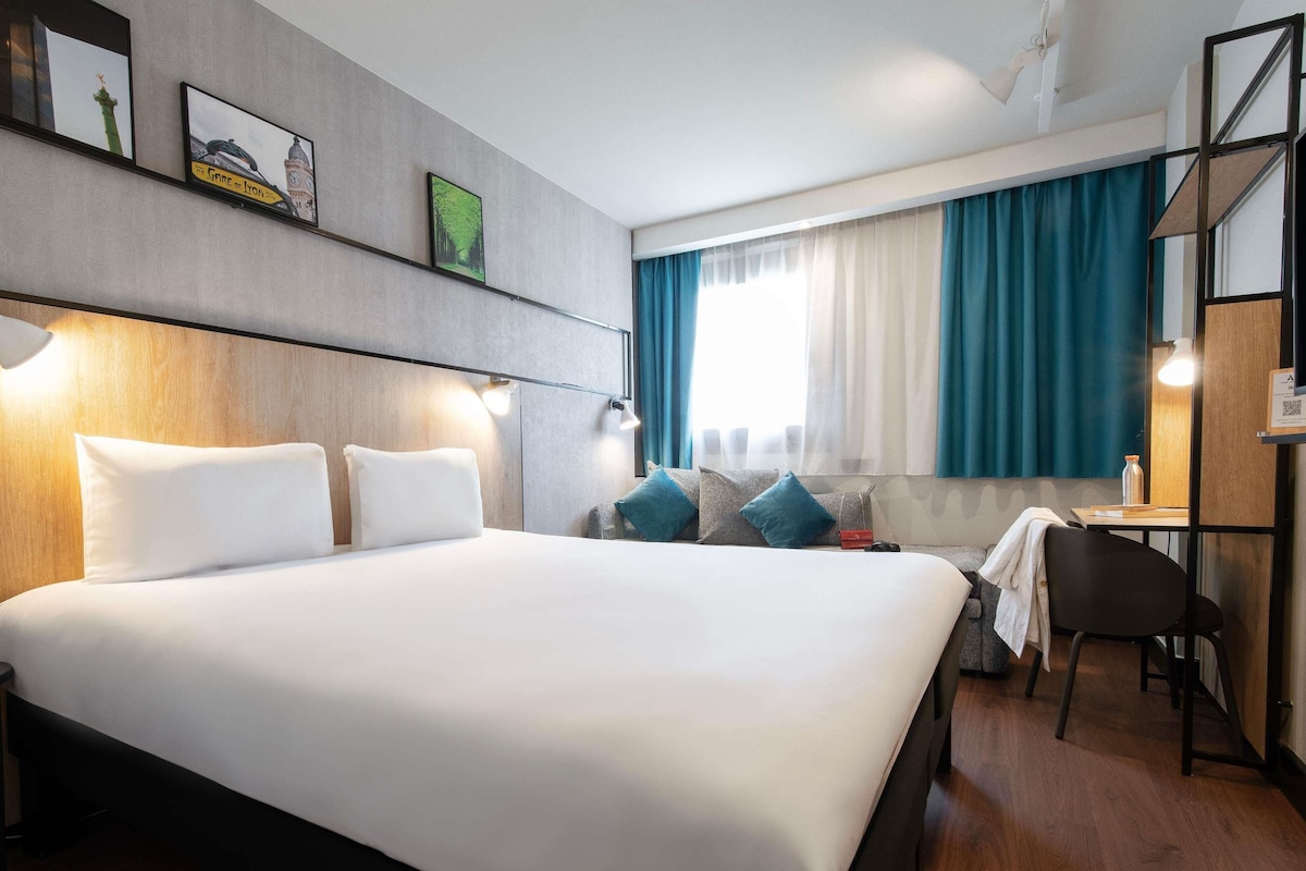 Een moderne hotelkamer met een groot bed, een bureau en een stoel. Er zijn twee ramen met gordijnen, en de kamer heeft een houten vloer.
