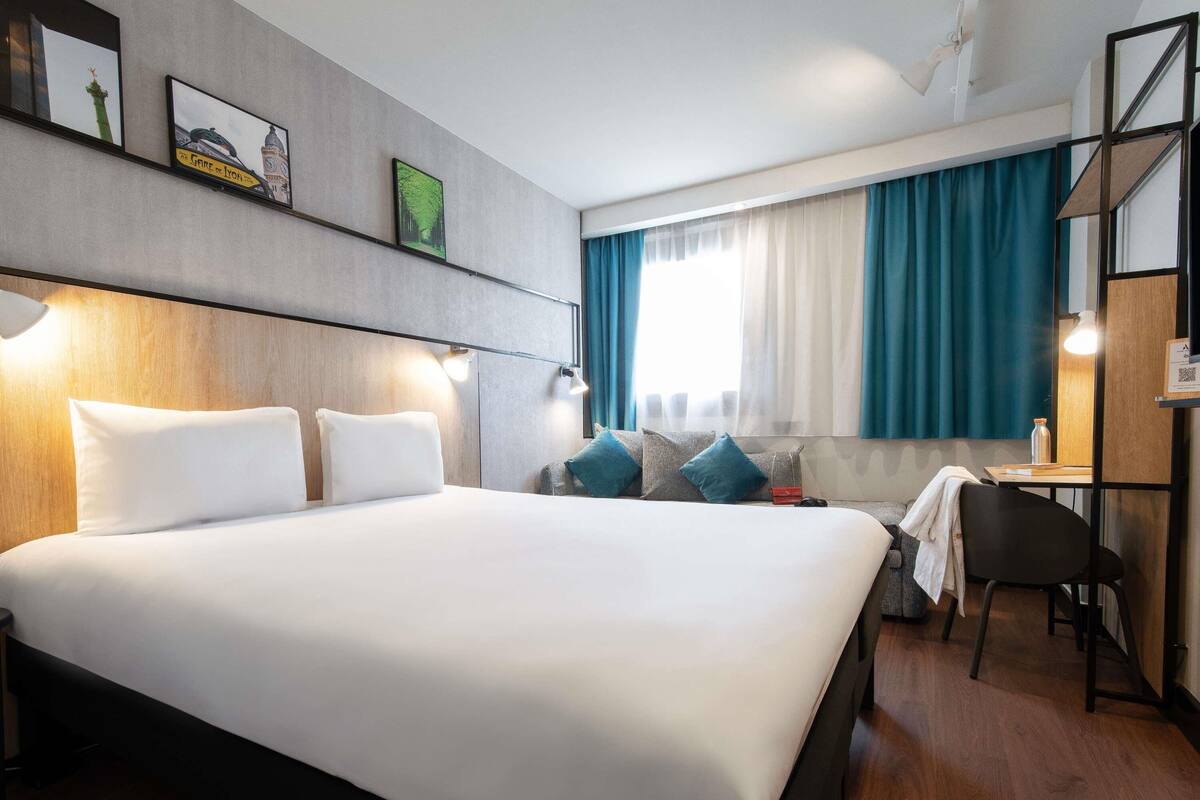 Een moderne hotelkamer met een groot bed, een bureau en een stoel. Er zijn twee ramen met gordijnen, en de kamer heeft een houten vloer.