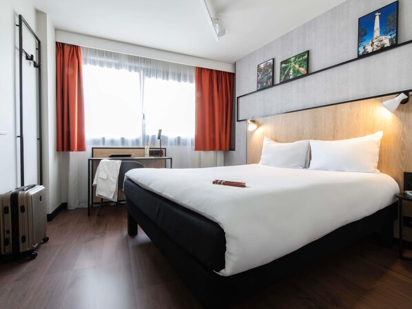 Una habitación de hotel con cama, escritorio, silla, maleta y ventana con cortinas.